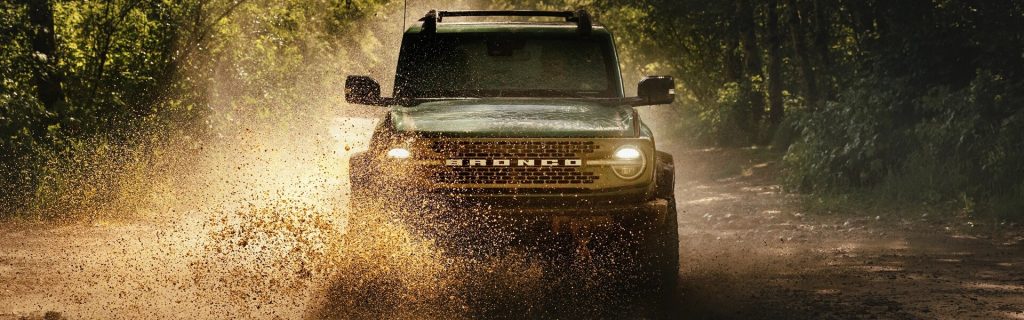 2025 Ford Bronco driving through a muddy forest trail, splashing water and dirt into the air, highlighting its rugged off-road capability and bold front grille design