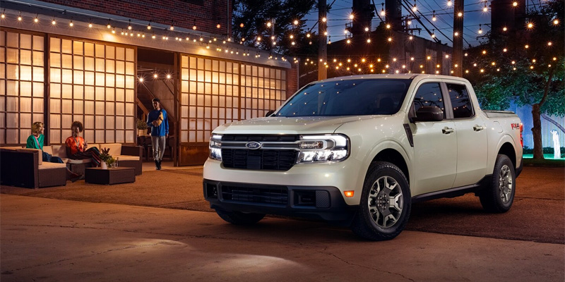 Front three-quarter view of a white Ford Maverick parked outdoors at night under string lights near a modern building with people seated in the background.